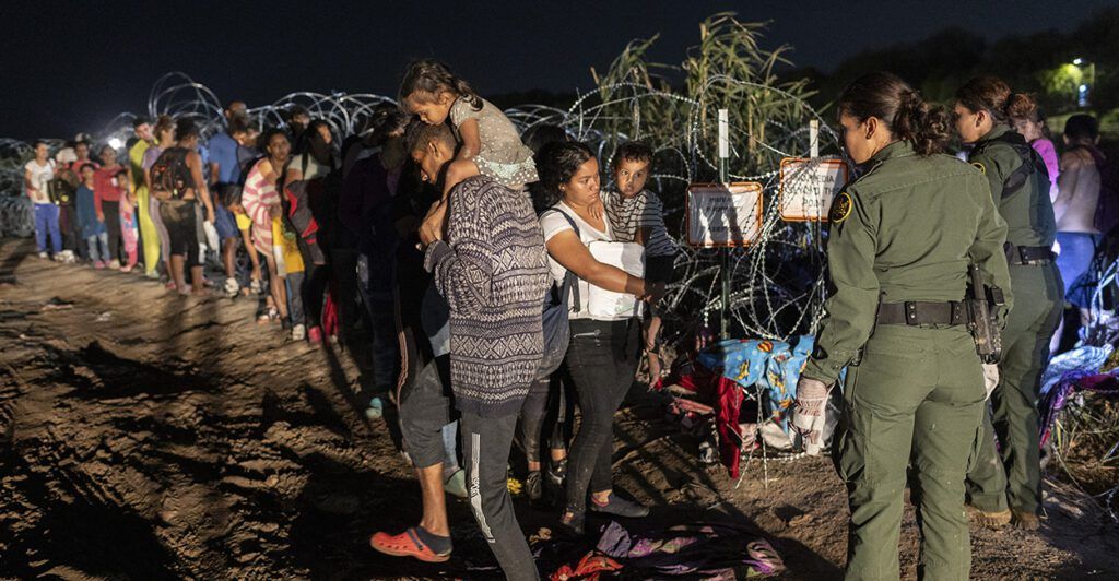 Migrants wait in line next to razor wire in the dark.