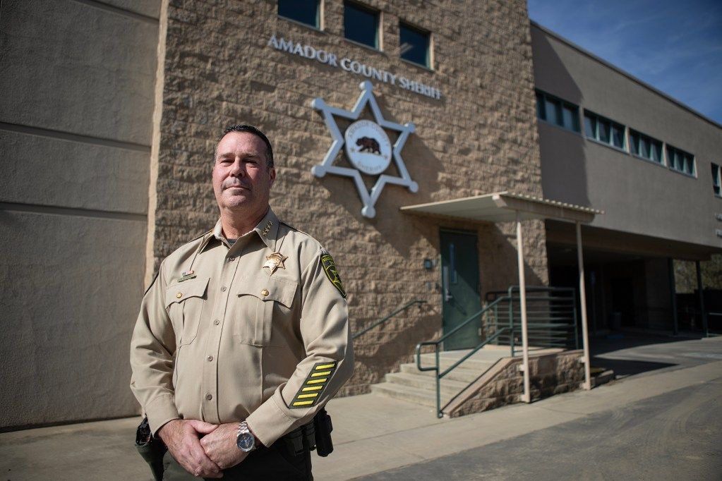 A uniformed officer stands outside the Amador County Sheriff’s Office, with a large sheriff’s badge emblem mounted on the brick wall behind them. The person has a confident expression, hands clasped in front, and is dressed in a tan uniform adorned with insignia and stripes on the sleeves. The building entrance, with a staircase and railing, is visible in the background under a clear blue sky.