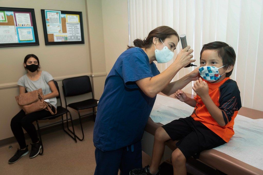 A healthcare worker in blue scrubs and a white mask examines a child's ear with an otoscope in a medical office. The child, wearing an orange shirt and a patterned face mask, sits on an examination table, slightly adjusting their mask. In the background, another person wearing a black mask and holding a brown purse sits on a chair, observing the examination. Educational posters are displayed on the beige walls.