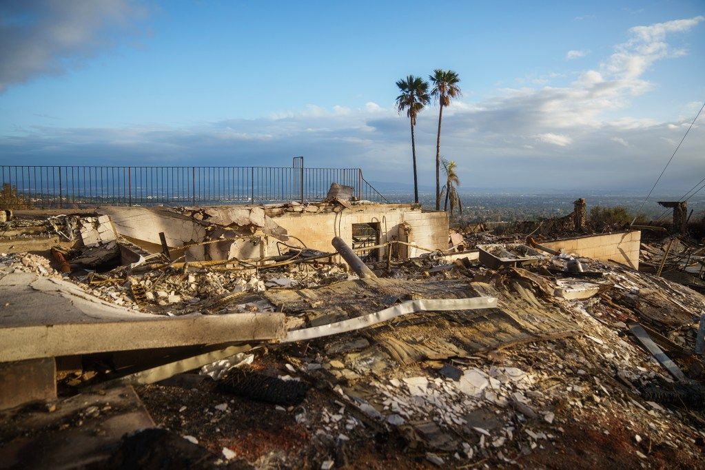 he burned remains of a hillside home with collapsed walls and scattered debris, overlooking a cityscape in the distance. A few scorched palm trees and a metal fence stand behind the ruins under a partly cloudy sky.