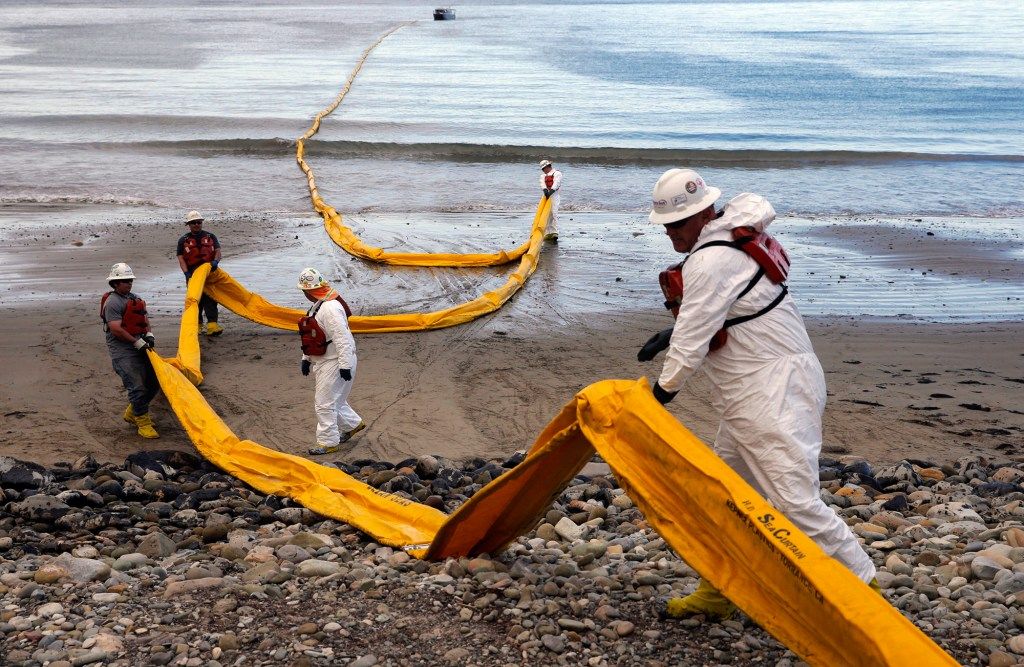 Cleanup workers in protective gear and life vests deploy a long yellow containment boom on a beach following an oil spill. The boom stretches from the rocky shore into the water, with a boat visible in the distance.