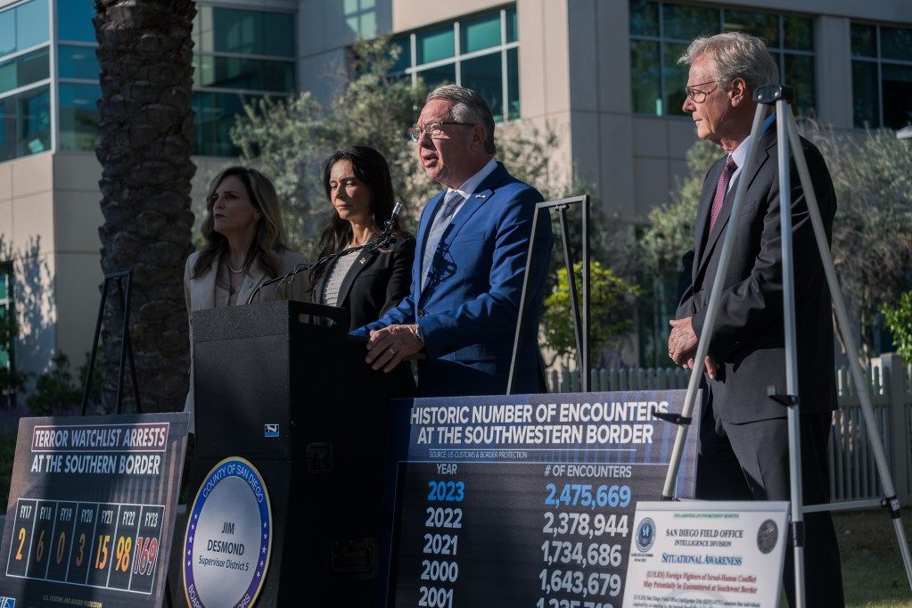 A side view of four people standing in front posters and a dais during a press conference. The person in the center has white hair and wears glasses and a bright blue suit.