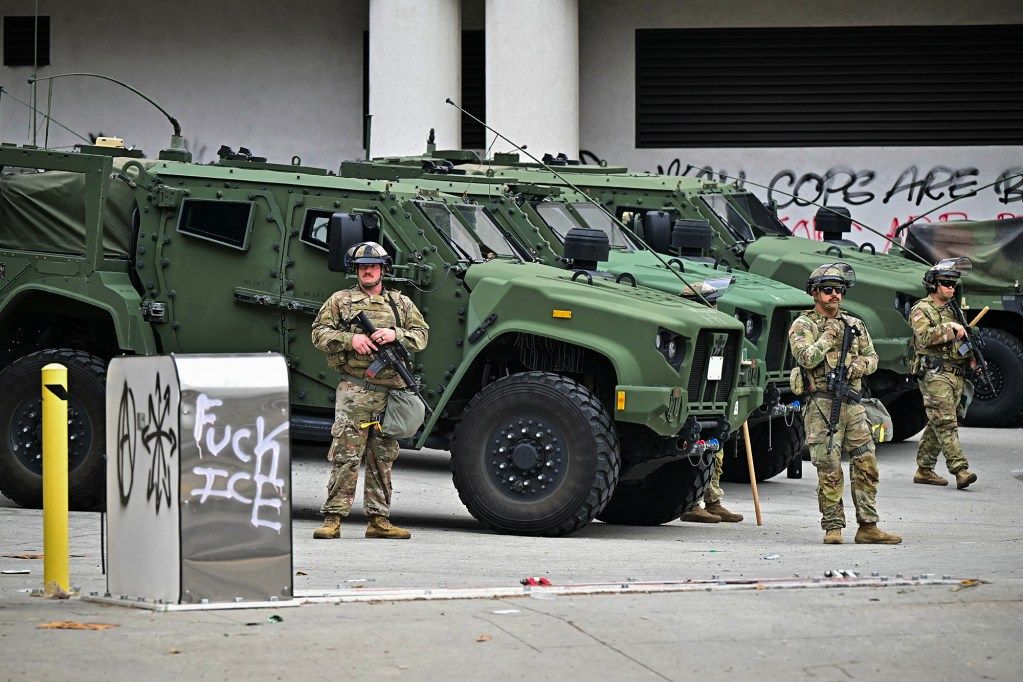 National Guard soldiers in camouflage uniforms and helmets stand guard with rifles in front of a row of armored green military vehicles in an urban area. Behind them, graffiti on a wall includes anti-police messages, and a nearby metal bin is tagged with
