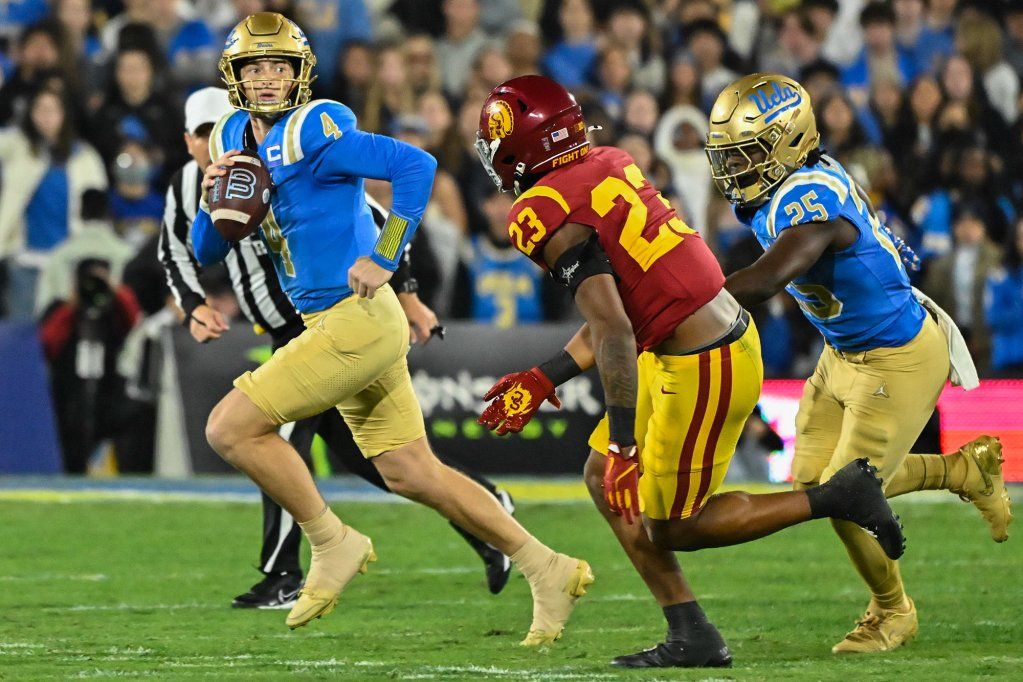 A football quarterback, dressed in a blue and gold uniform, runs while holding a football as a player from the opposing team tried to block them while also being chased by another player.