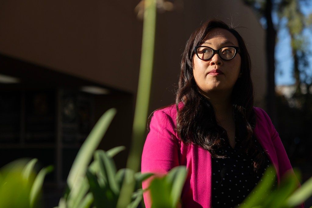 A person in glasses and a jacket stares into the distance, seated next to vegetation. At the bottom of the frame are pieces of a plate poking into it, with one stem passing through the center. The person has rays of golden sunlight shining through one side of their body.
